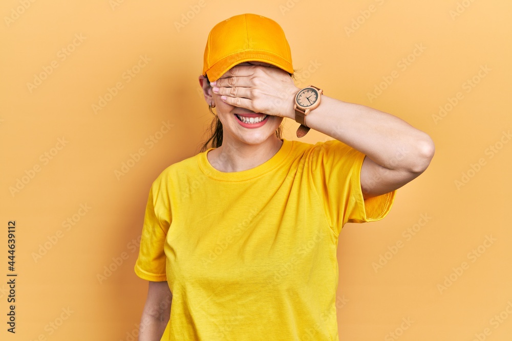 Young hispanic woman wearing delivery uniform and cap smiling and laughing with hand on face covering eyes for surprise. blind concept.