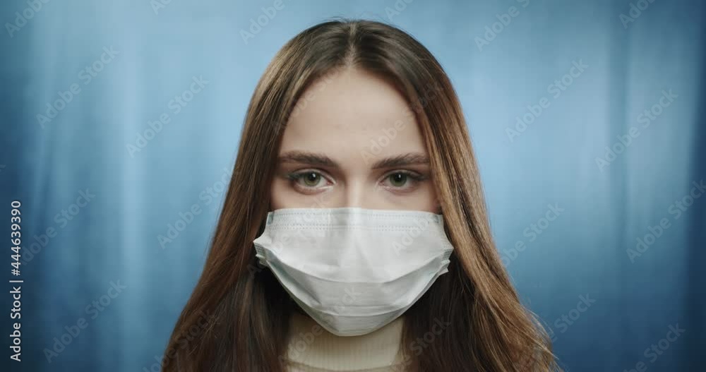 Covid-19, pandemic and quarantine concept. Close-up of serious teenager student woman wearing medical protective mask during coronavirus, standing against blue background. Covid-19, pandemic and quarantine concept. Close-up of serious teenager student woman wearing medical protective mask during coronavirus, standing against blue background.