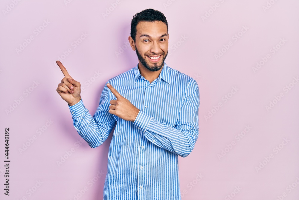 Hispanic man with beard wearing casual blue shirt smiling and looking at the camera pointing with two hands and fingers to the side. Hispanic man with beard wearing casual blue shirt smiling and looking at the camera pointing with two hands and fingers to the side.