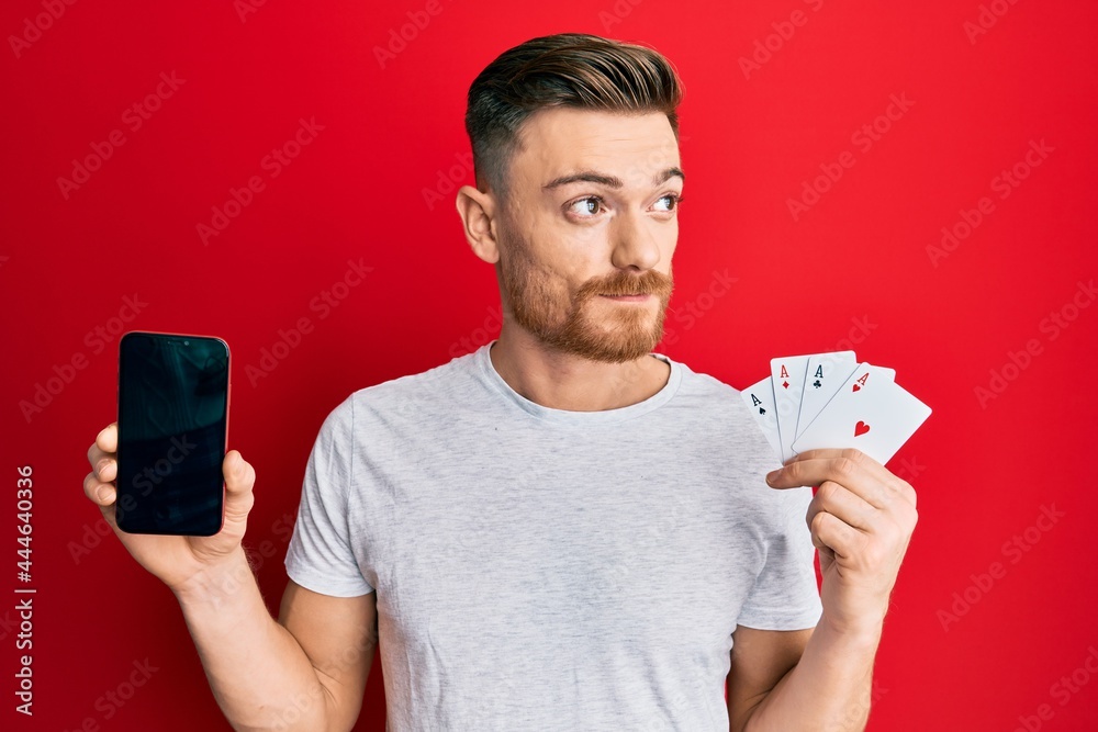 Young redhead man holding smartphone and casino card smiling looking to the side and staring away thinking. Young redhead man holding smartphone and casino card smiling looking to the side and staring away thinking.