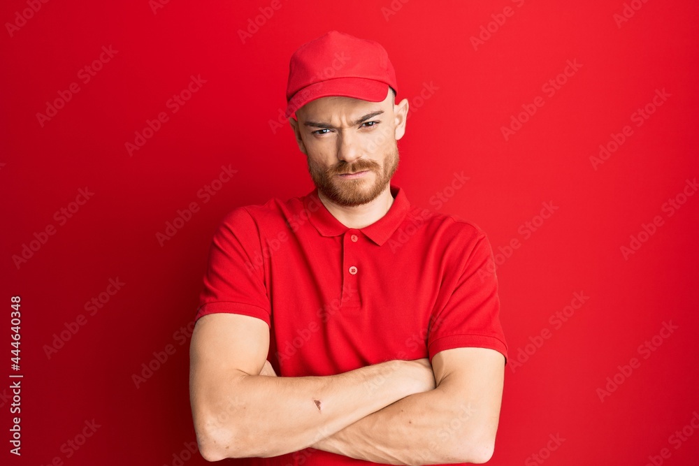 Young redhead man wearing delivery uniform and cap skeptic and nervous, disapproving expression on face with crossed arms. negative person.