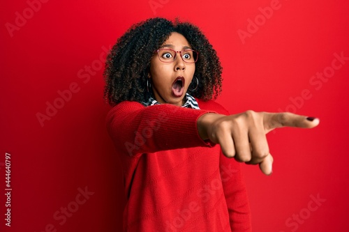 Beautiful african american woman with afro hair wearing sweater and glasses pointing with finger surprised ahead, open mouth amazed expression, something on the front