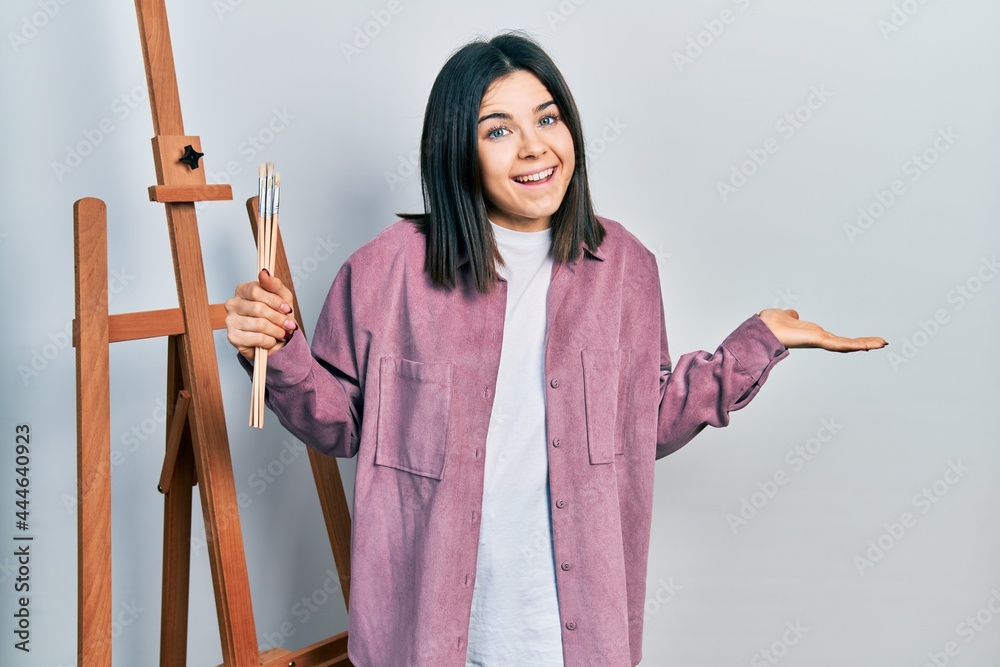 Young brunette woman painter sitting at art studio holding art word celebrating achievement with happy smile and winner expression with raised hand Young brunette woman painter sitting at art studio holding art word celebrating achievement with happy smile and winner expression with raised hand