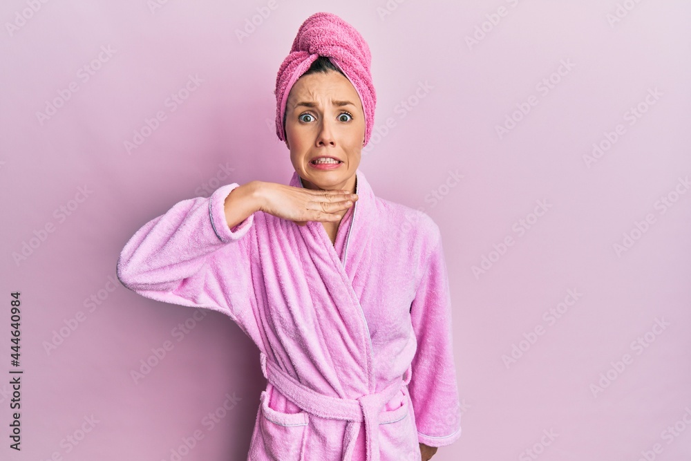Young blonde woman wearing shower towel cap and bathrobe cutting throat with hand as knife, threaten aggression with furious violence Young blonde woman wearing shower towel cap and bathrobe cutting throat with hand as knife, threaten aggression with furious violence