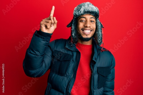 Young african american man with beard wearing winter hat and coat pointing finger up with successful idea. exited and happy. number one. photo