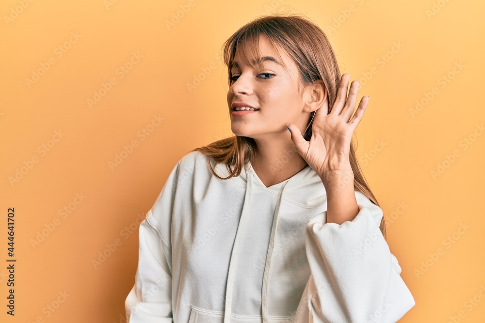 Teenager caucasian girl wearing casual sweatshirt smiling with hand over ear listening and hearing to rumor or gossip. deafness concept. Teenager caucasian girl wearing casual sweatshirt smiling with hand over ear listening and hearing to rumor or gossip. deafness concept.