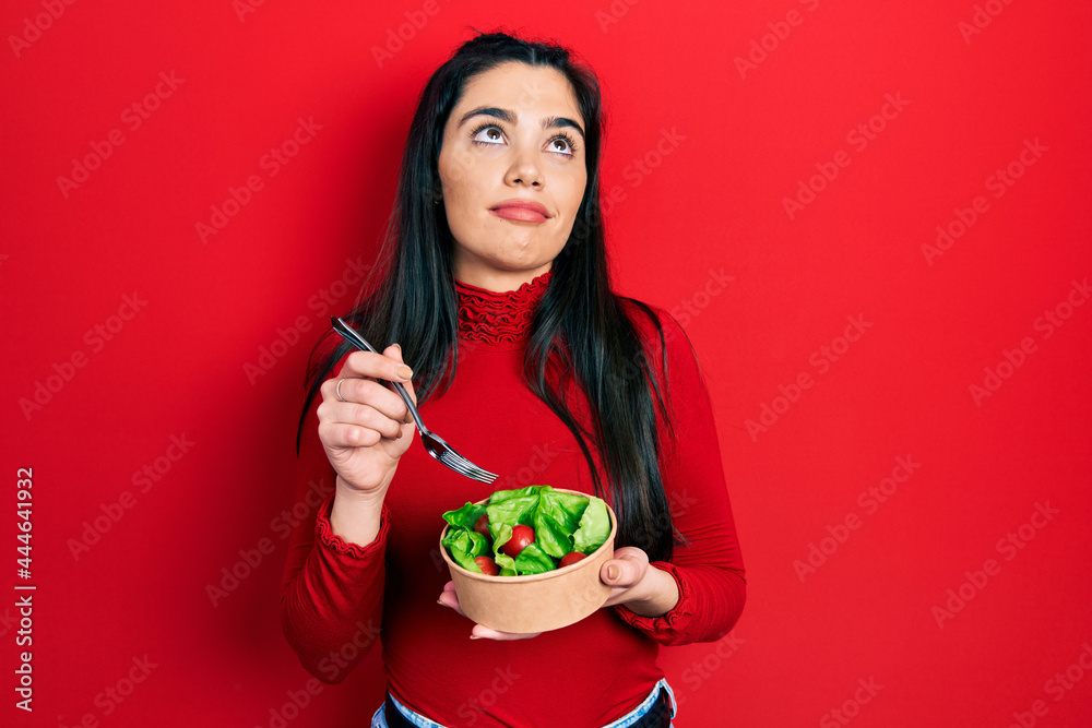 Young hispanic girl eating salad smiling looking to the side and staring away thinking.