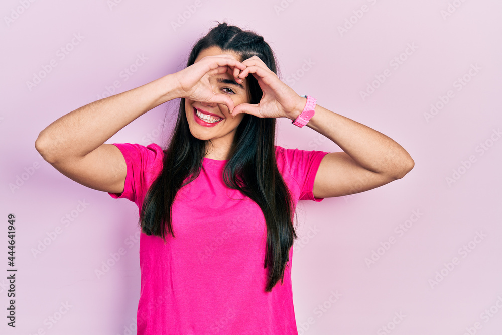 Young hispanic girl wearing casual pink t shirt doing heart shape with hand and fingers smiling looking through sign Young hispanic girl wearing casual pink t shirt doing heart shape with hand and fingers smiling looking through sign