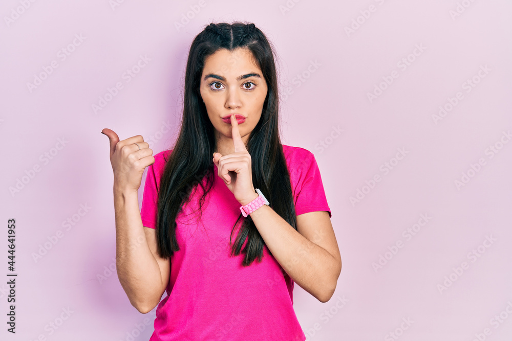 Young hispanic girl wearing casual pink t shirt asking to be quiet with finger on lips pointing with hand to the side. silence and secret concept. Young hispanic girl wearing casual pink t shirt asking to be quiet with finger on lips pointing with hand to the side. silence and secret concept.