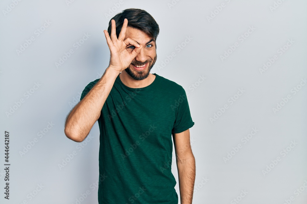 Young hispanic man wearing casual white tshirt doing ok gesture with hand smiling, eye looking through fingers with happy face. Young hispanic man wearing casual white tshirt doing ok gesture with hand smiling, eye looking through fingers with happy face.