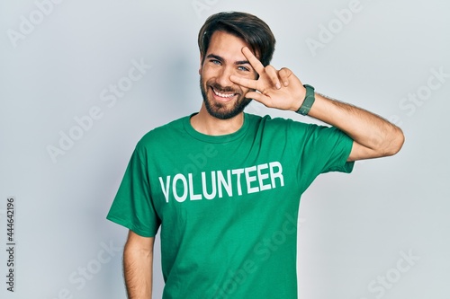 Young hispanic man wearing volunteer t shirt doing peace symbol with fingers over face, smiling cheerful showing victory