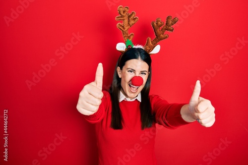 Young hispanic woman wearing deer christmas hat and red nose approving doing positive gesture with hand  thumbs up smiling and happy for success. winner gesture.