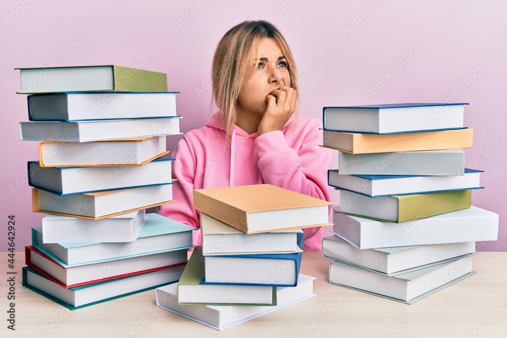Young caucasian woman sitting on the table with books looking stressed and nervous with hands on mouth biting nails. anxiety problem. Young caucasian woman sitting on the table with books looking stressed and nervous with hands on mouth biting nails. anxiety problem.