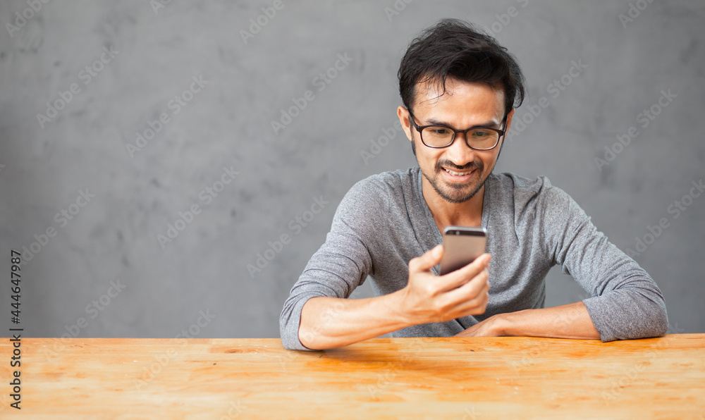 happy young man smiling and using mobile phone with wooden table and concrete wall background and copy space happy young man smiling and using mobile phone with wooden table and concrete wall background and copy space