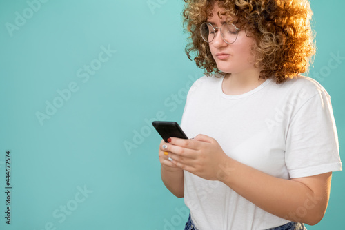 Internet haters. Fat shaming. Obesity problem. Body positive. Sad frowning overweight young woman with curly hair reading message on phone isolated on blue copy space background.