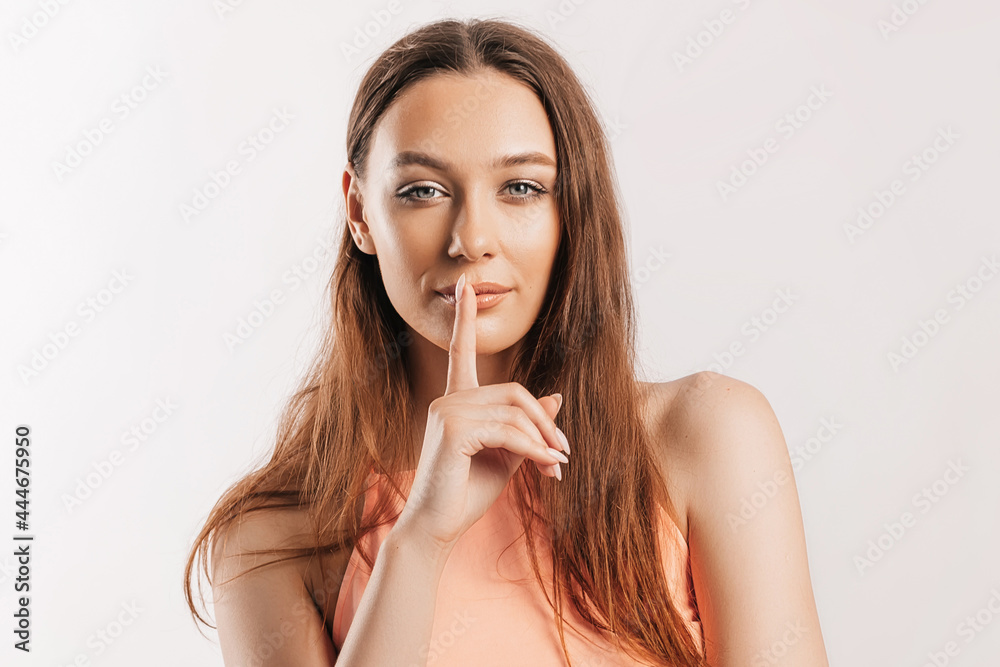 Shh gesture. Young beautiful serious girl holding a finger to her lips on a white isolated background. A woman asks to be silent, a place for advertising. Positive brunette in orange top Shh gesture. Young beautiful serious girl holding a finger to her lips on a white isolated background. A woman asks to be silent, a place for advertising. Positive brunette in orange top