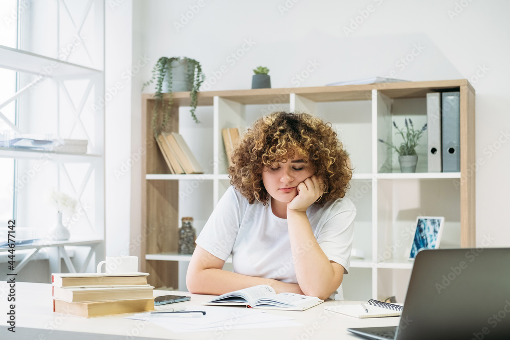 Demotivated student. Exam study. Home learning. Bored overweight young woman with curly hair resting head on hand reading book in light modern interior room. Demotivated student. Exam study. Home learning. Bored overweight young woman with curly hair resting head on hand reading book in light modern interior room.