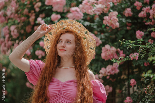 Beautiful happy smiling redhead woman looking up. Model with long natural curly hair  wearing trendy summer pink blouse, straw hat,  posing in blooming rose garden. Outdoor lifestyle portrait