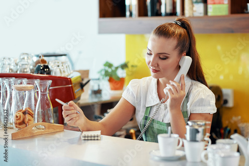 Young restaurant waitress taking order by phone and writing details in notepad