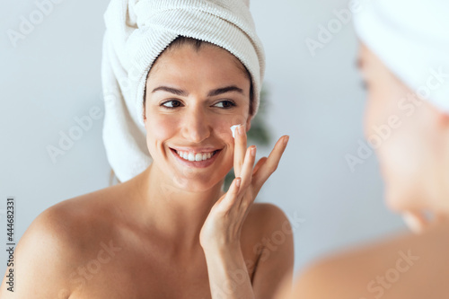 Beautiful young woman caring of her skin while putting on cream looking at mirror at home.