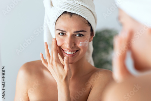 Beautiful young woman caring of her skin while putting on cream looking at mirror at home.