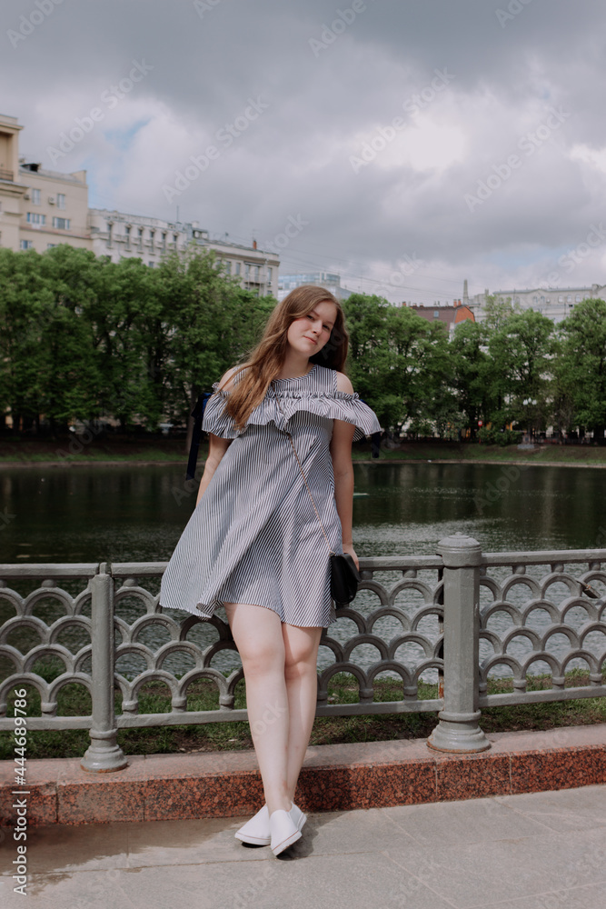 Smiling young woman in dress standing alone among street, leaning on fence in front of pond. looking at camera. Having fun outside. Urban style and street fashion. Sumeer casual outfit