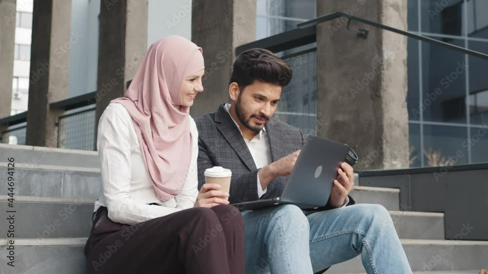 Woman in hijab and man in suit sitting on stairs near office building and discussing common project. Arabian partners using modern laptop for work outdoors. Two arabian colleagues sitting on stairs Woman in hijab and man in suit sitting on stairs near office building and discussing common project. Arabian partners using modern laptop for work outdoors. Two arabian colleagues sitting on stairs