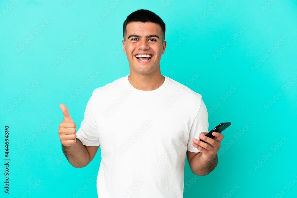 Young handsome man over isolated blue background using mobile phone while doing thumbs up Young handsome man over isolated blue background using mobile phone while doing thumbs up
