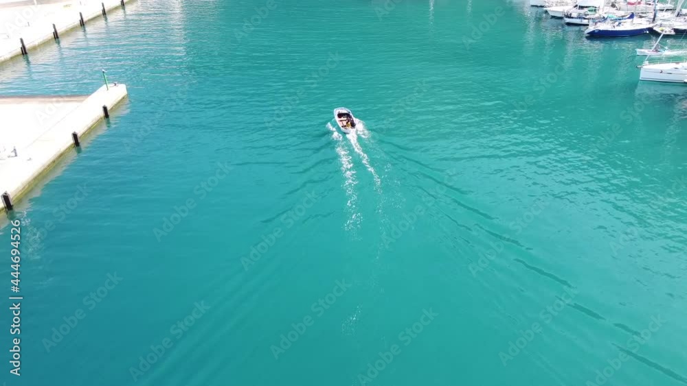 Following small sailboat navigating turquoise water on route leaving sunny marina aerial view from behind