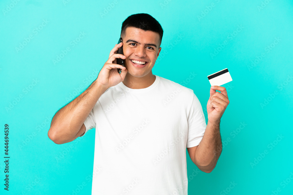 Young handsome man over isolated blue background keeping a conversation with the mobile phone and holding a credit card Young handsome man over isolated blue background keeping a conversation with the mobile phone and holding a credit card