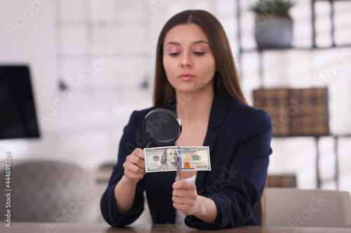 Expert authenticating 100 dollar banknote with magnifying glass at table in office, focus on hand. Fake money concept photo