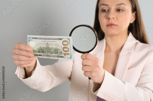 Expert authenticating 100 dollar banknote with magnifying glass against light grey background, focus on hand. Fake money concept photo