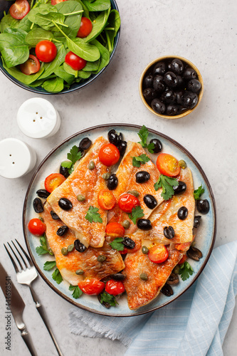 Perch fish fillets stew, italian style, with black olives, tomatoes and capers, with fresh parsley and fresh salad. On light grey background. Top view. Vertical image. photo