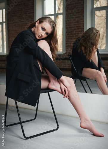 Studio fashion shot of attractive young woman. Portrait of beautiful girl dressed in black jacket (blazer). Lovely lady sitting in chair at the mirror