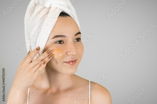 Beauty portrait of smiling woman in towel on head applying yellow nourishing mask on face on gray isolated background. Skin care concept. 