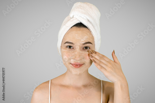 Happy young woman with bath towel on her head applies cleansing foam and smiles. Isolated on a gray background. High quality photo