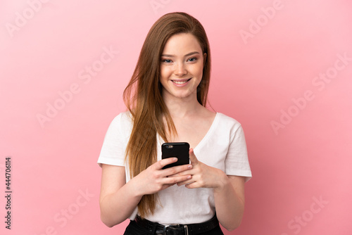 Teenager girl over isolated pink background looking at the camera and smiling while using the mobile