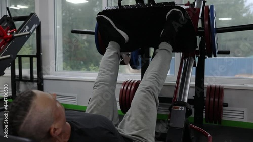 A man in sportswear, with tattoos, his hair partially shaved off, does a platform press with his legs.