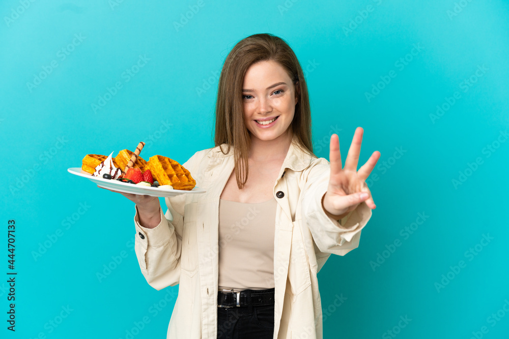 Teenager girl holding waffles over isolated blue background happy and counting three with fingers Teenager girl holding waffles over isolated blue background happy and counting three with fingers