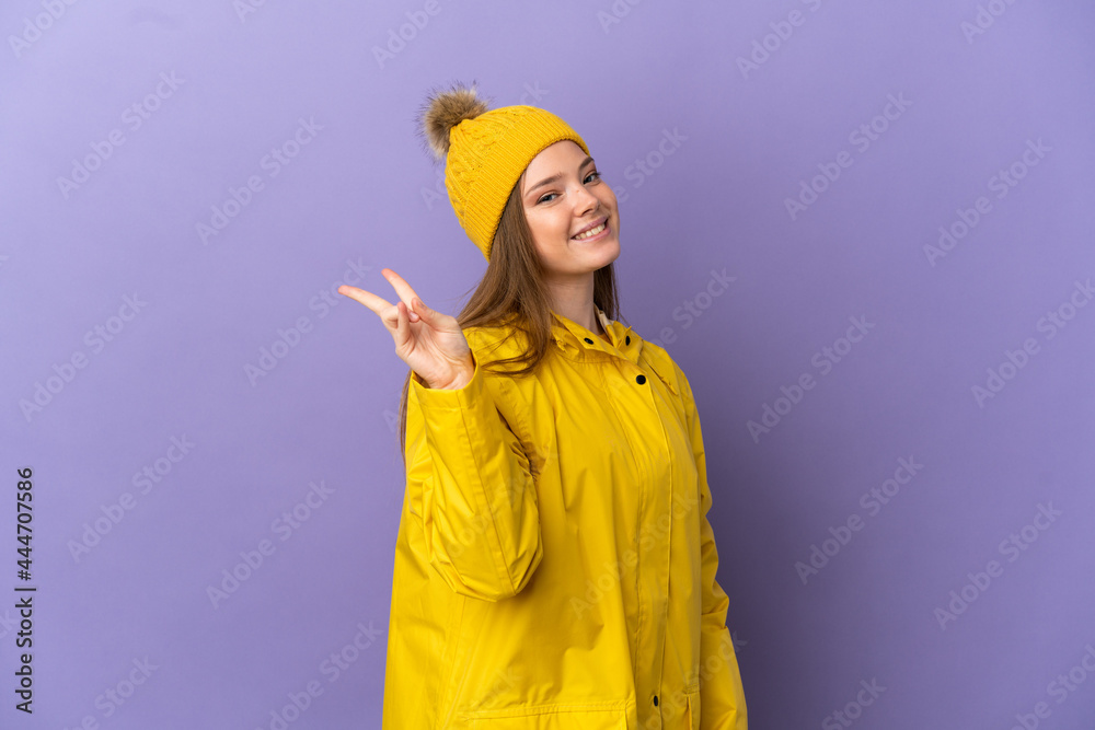 Teenager girl wearing a rainproof coat over isolated purple background smiling and showing victory sign Teenager girl wearing a rainproof coat over isolated purple background smiling and showing victory sign
