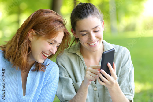Two happy friends laughing checking phone in a park