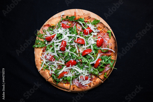 Pizza with tomato sauce, smoked pork muscles, mozzarella, arugula salad and cherry tomatoes on a black background.