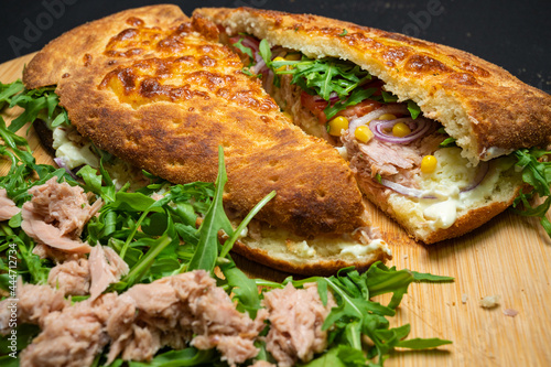 Tuna sandwich, cheese sauce, tomatoes, red onions, corn and arugula salad on a wooden plate on a black background.