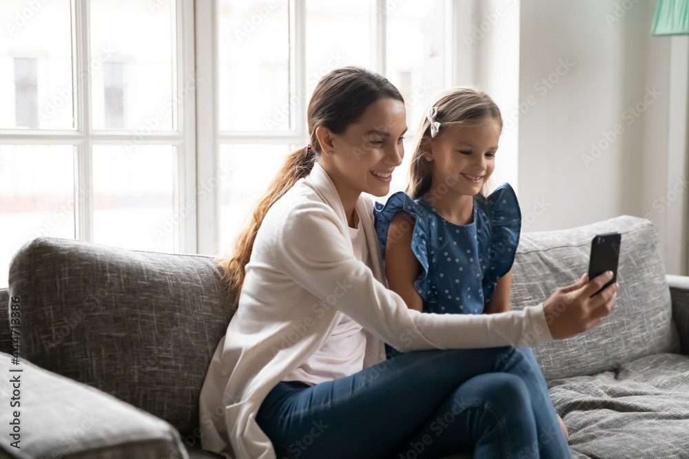 Happy young Caucasian mother and little daughter relax on sofa have webcam virtual digital event on cellphone. Smiling mom and small girl child talk speak on video call on modern smartphone. Happy young Caucasian mother and little daughter relax on sofa have webcam virtual digital event on cellphone. Smiling mom and small girl child talk speak on video call on modern smartphone.