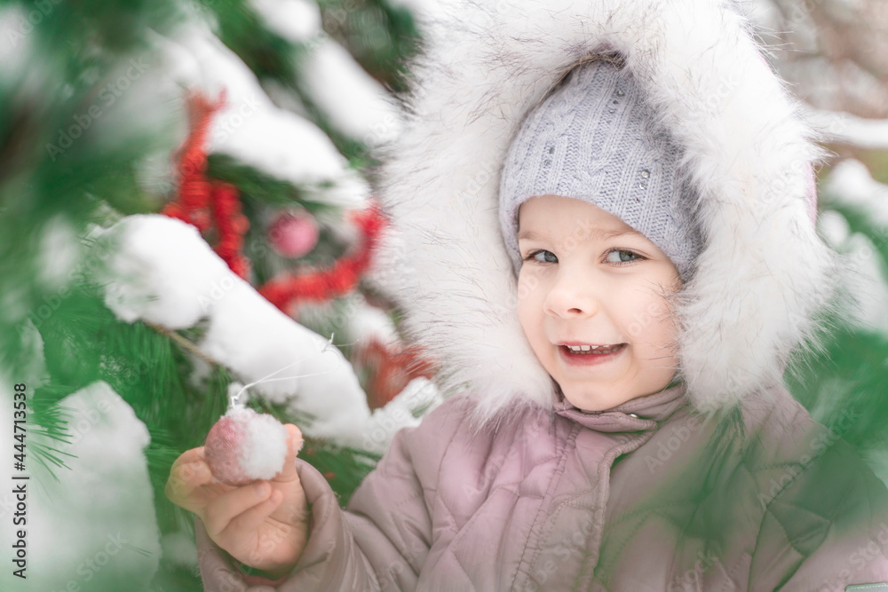 A close up portrait of a little cute smiling girl in a knitted cap standing near the decorated Christmas tree on a frosty winter day outdoor.