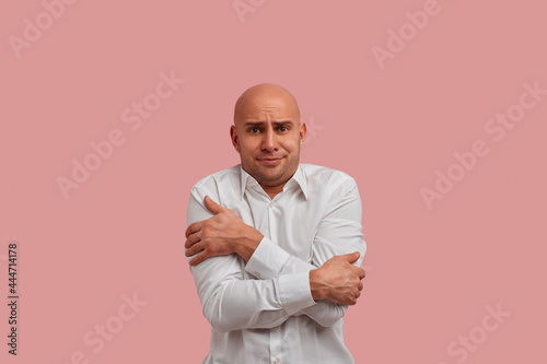 Make it warmer, please. Frozen bald man with bristle trembles from cold in office. Dissatisfied with the cold from the air conditioner. Dressed in white shirt. Isolated over pink background.