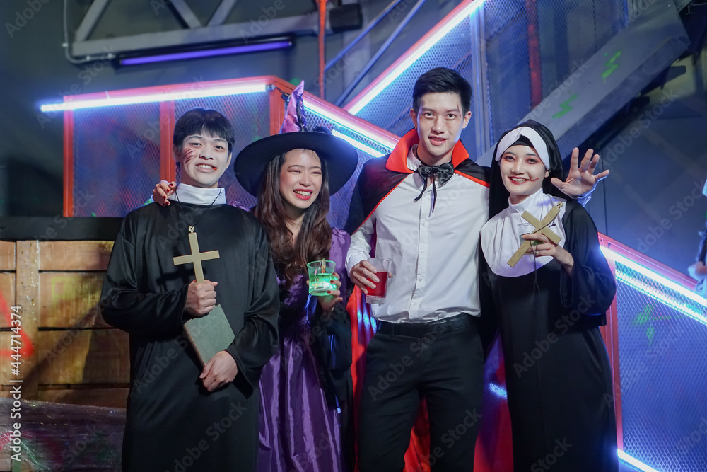 Group of young Asian friends dressed as witch, priest, vampire and nun makeup face with blood and wounds smiling and holding drinks at Halloween party and having fun at night club