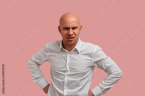 Portrait of crazy bald man with bristle makes angry grimace, showing tooth grin, hold fists on belt. Frowned, dissatisfied with the act of his children. Isolated over pink background.