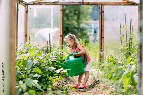 A cheerful baby with a watering can waters the plants in the greenhouse, helps to take care of them. Childhood, mother's assistant, plant care