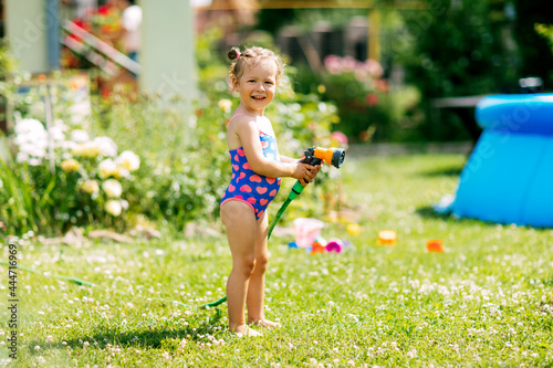 Charming baby girl waters plants with a hose in the garden in the backyard of the house on a sunny summer day